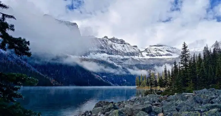Lago rodeado de bosque y montañas nevadas en Alaska con nubes bajas sobre el paisaje