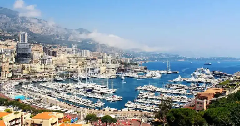 Vista panorámica del puerto de Mónaco con yates, edificios, montañas y el mar Mediterráneo en la Costa Azul.