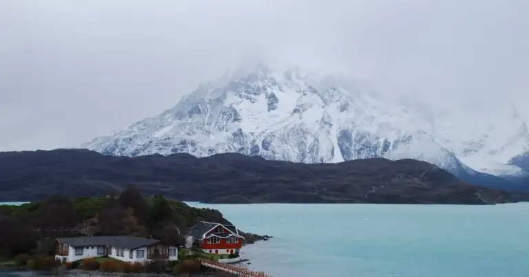 Lago de color turquesa con montañas nevadas y cabañas en la Patagonia chilena, una imagen que representa los paisajes ideales para unas vacaciones de invierno en Chile.