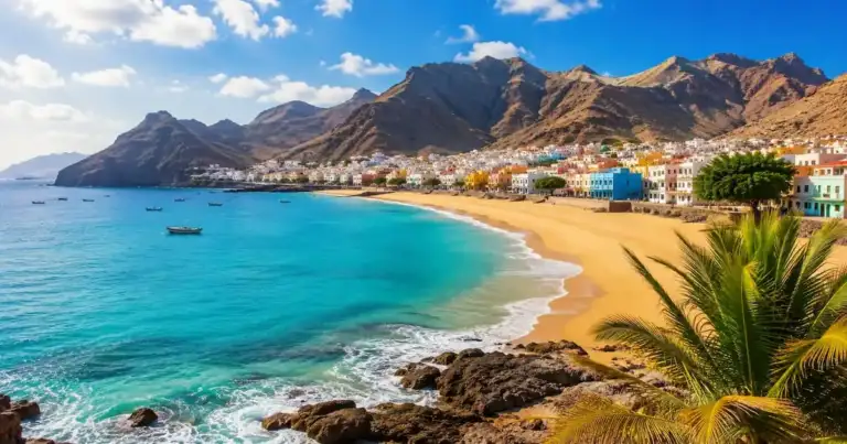 Vista panorámica de una playa de arena dorada en Cabo Verde, con mar turquesa, un pueblo costero de casas coloridas y montañas al fondo.