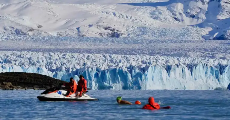 Nadador compitiendo frente al glaciar Perito Moreno con apoyo de seguridad en moto de agua durante una prueba en aguas heladas.
