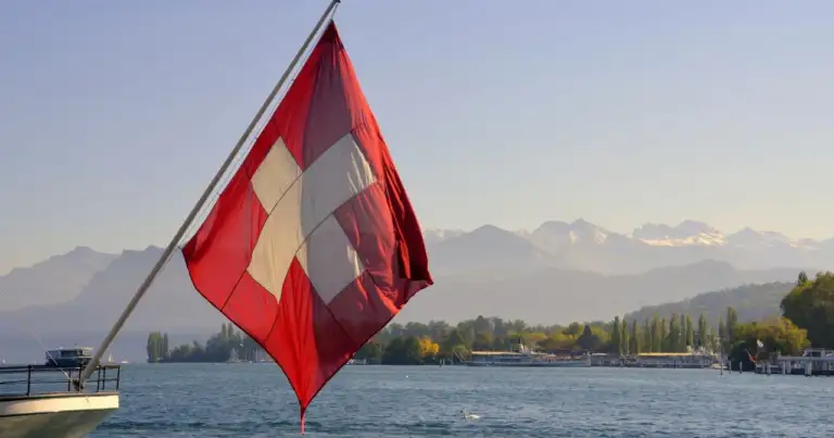 Bandera de Suiza en un barco sobre un lago, con montañas alpinas al fondo, imagen relacionada con dónde queda Suiza.