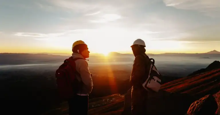 Dos viajeros con mochilas contemplando el amanecer desde una zona de montaña
