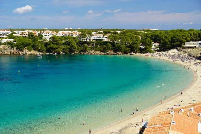 Menorca con niños: playas y vacaciones en familia Playa de aguas turquesas rodeada de pinares y casas blancas en Menorca, Islas Baleares