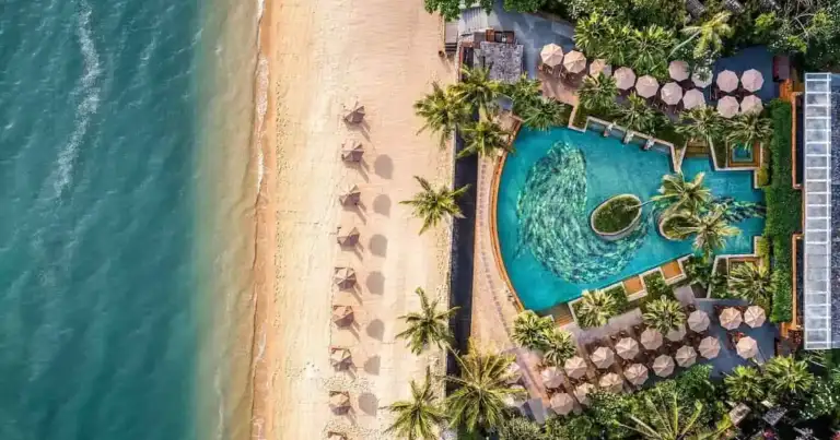 Vista aérea de un resort frente a la playa con piscina, palmeras y sombrillas junto al mar